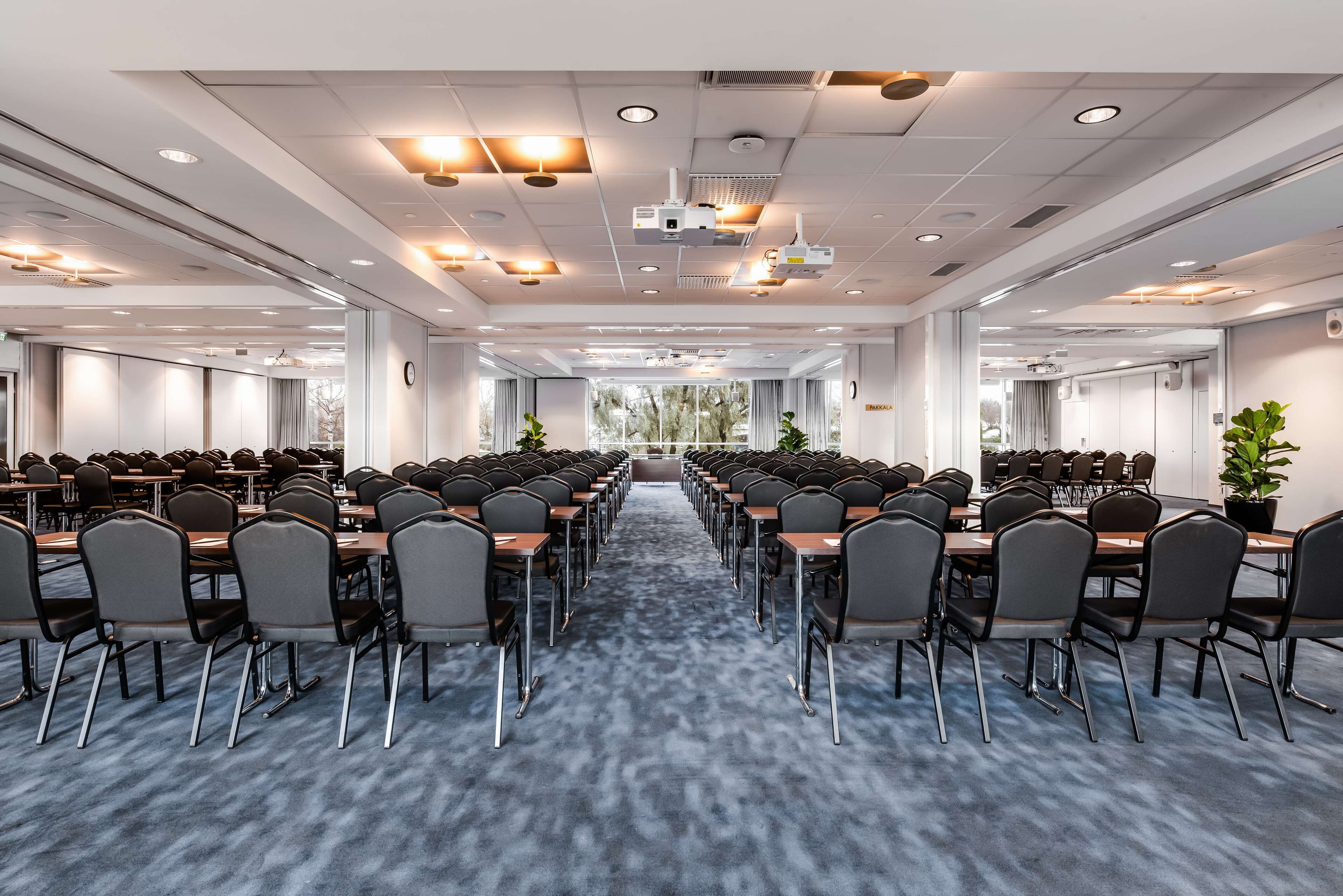 A conference room with rows of tables and chairs, large windows, and a carpeted floor.
