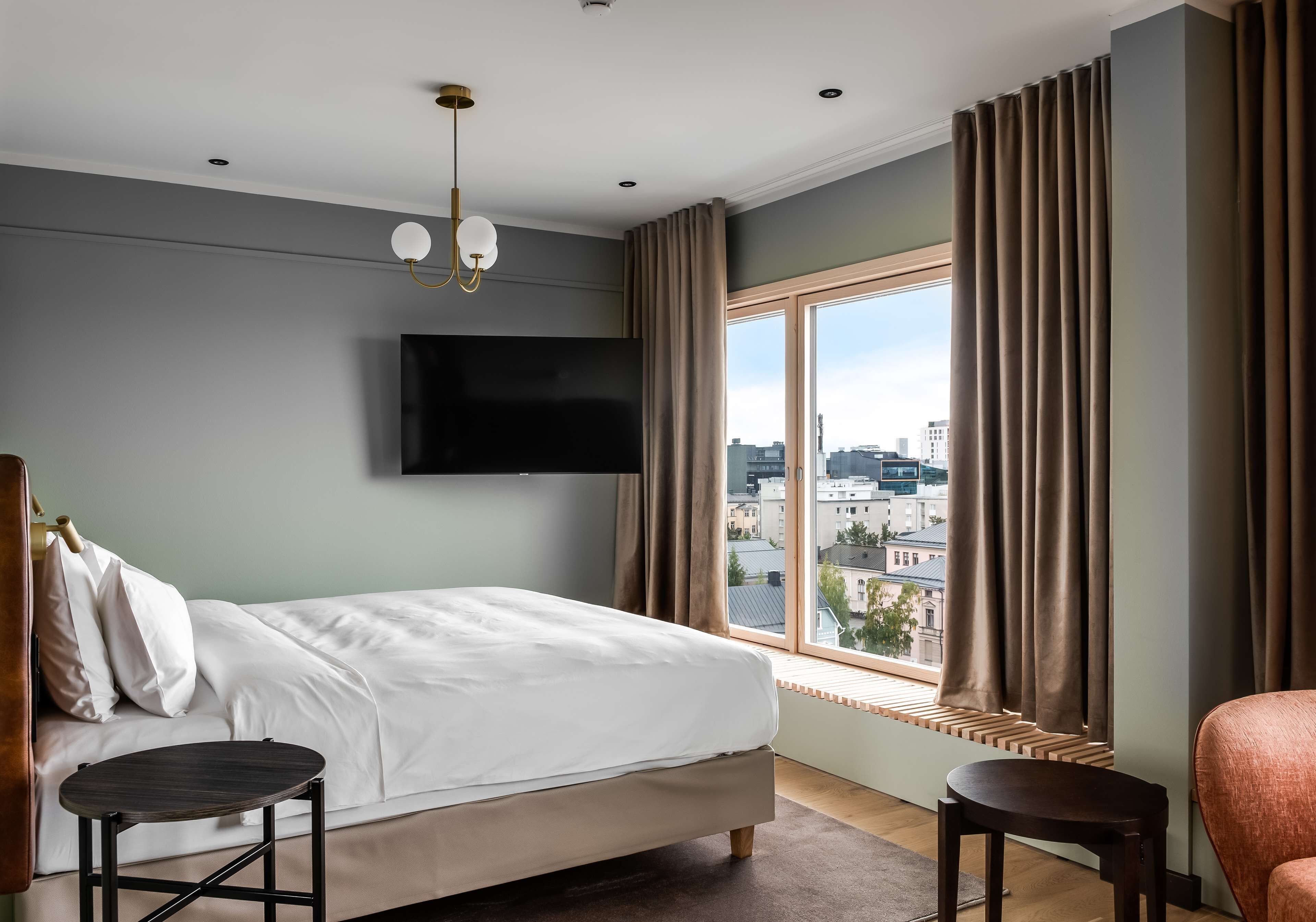 A hotel room with a bed, a TV mounted on the wall, a window with curtains, and a view of the cityscape.