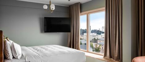 A hotel room with a bed, a TV mounted on the wall, a window with curtains, and a view of the cityscape.