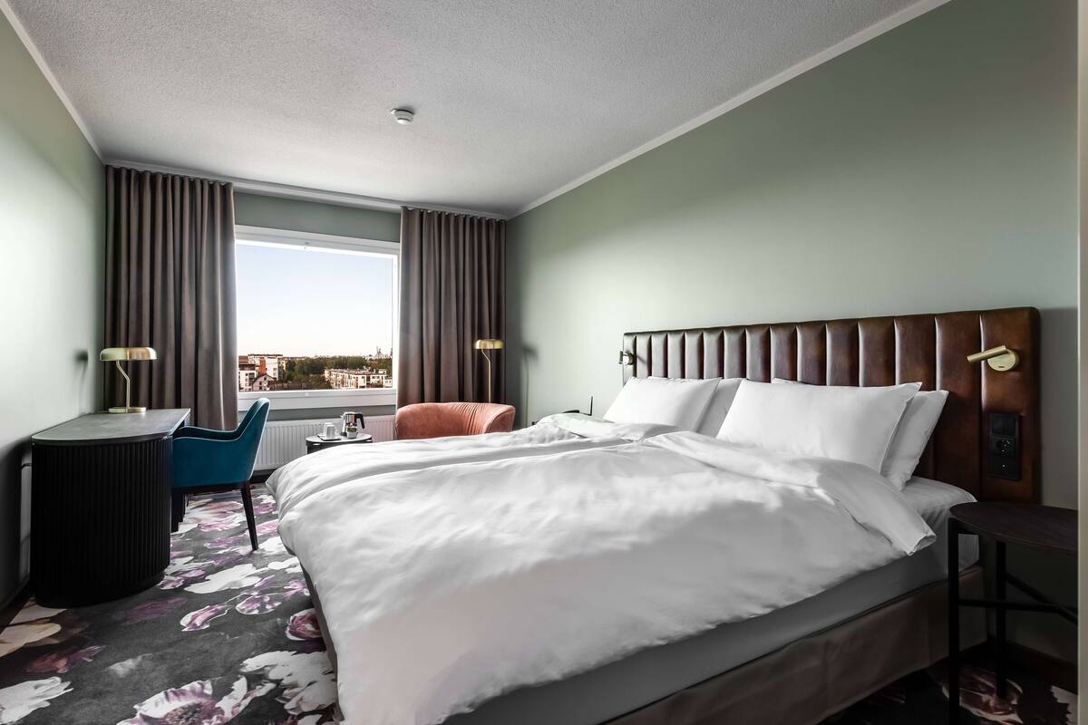 A hotel room with a large bed, a desk, a chair, and a view of the city.