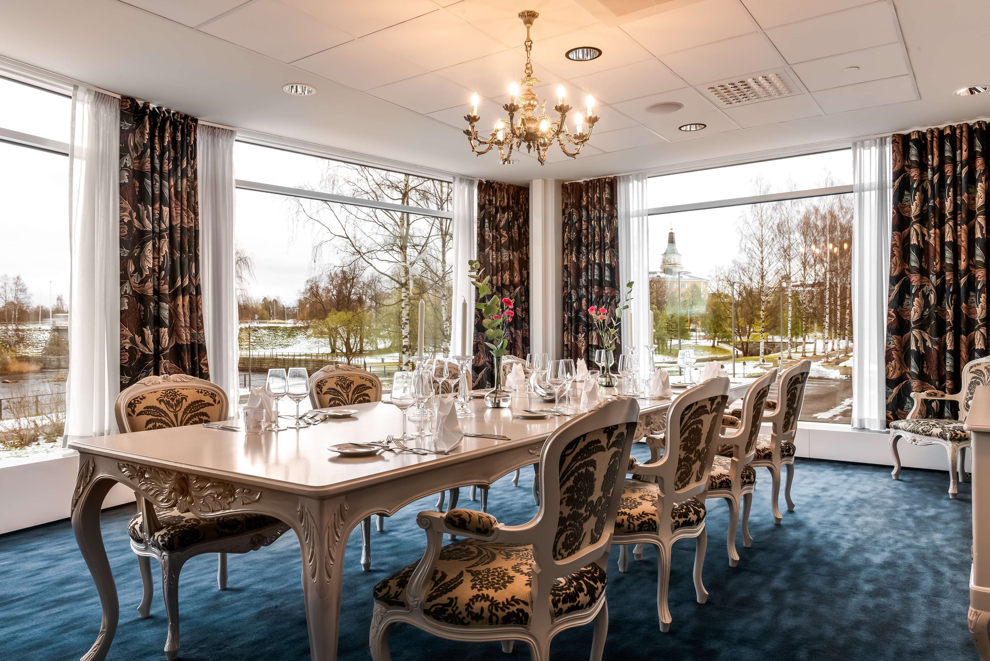 A dining room with a long table set for six, surrounded by ornate chairs, large windows with curtains, and a chandelier.