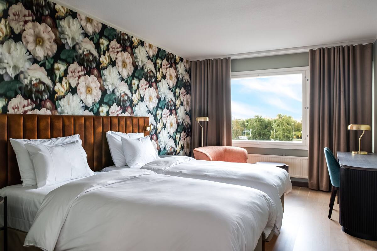 A hotel room with two beds, a large floral wallpaper, a view of trees, and a desk with a lamp.