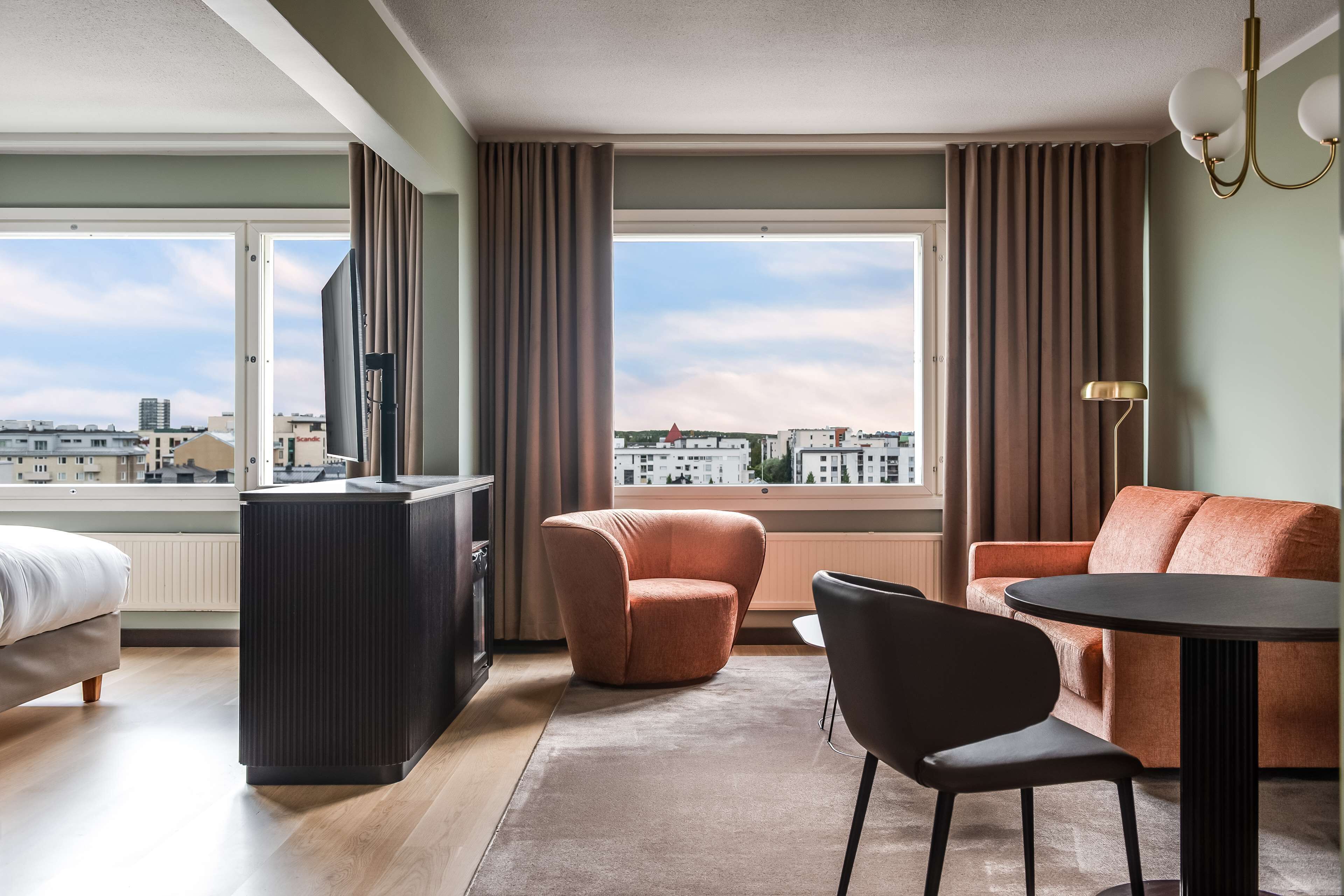 A modern hotel room with a bed, a desk, a sofa, and a view of the city.