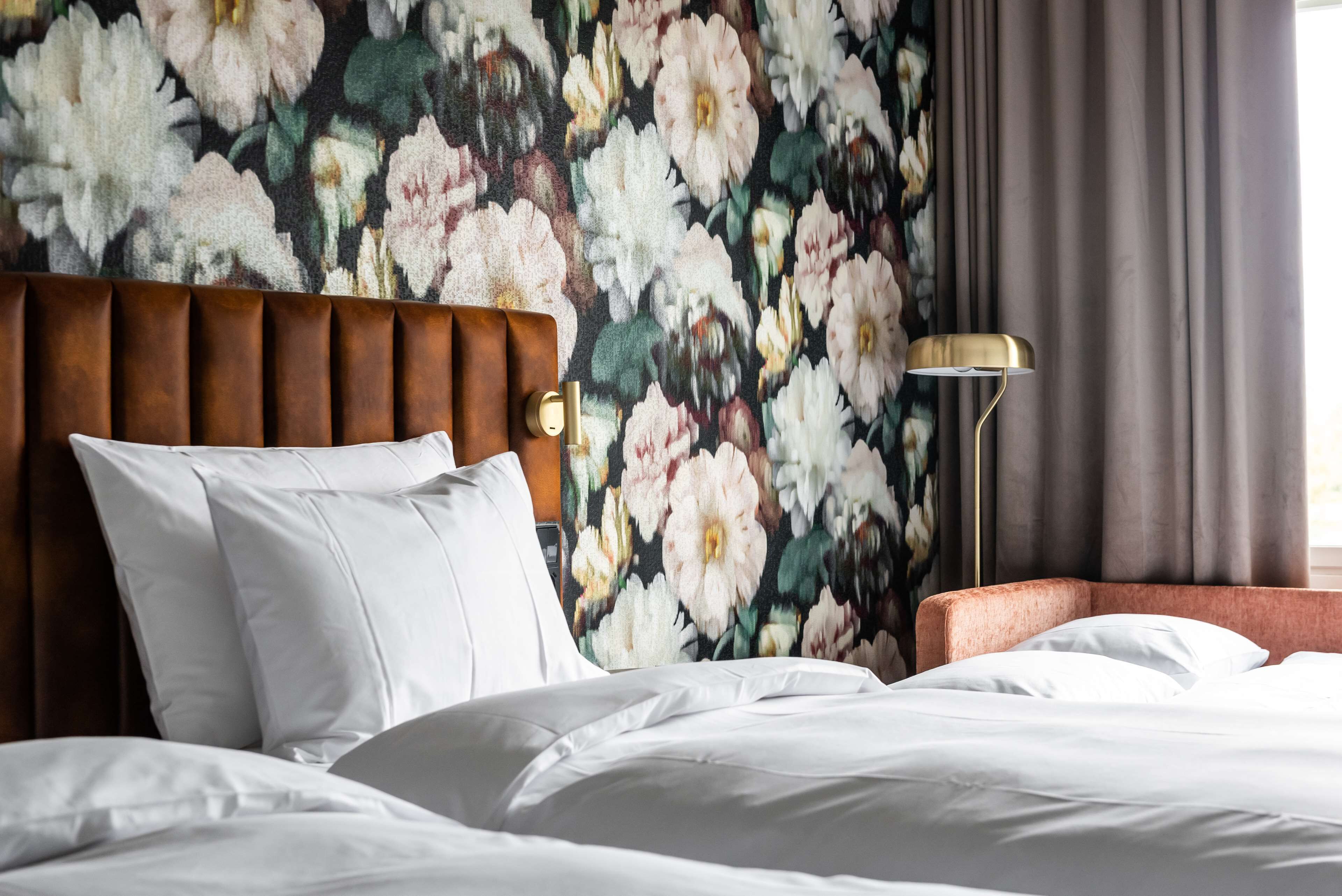 A neatly made bed with white linens, a brown upholstered headboard, and a floral-patterned wall behind it.