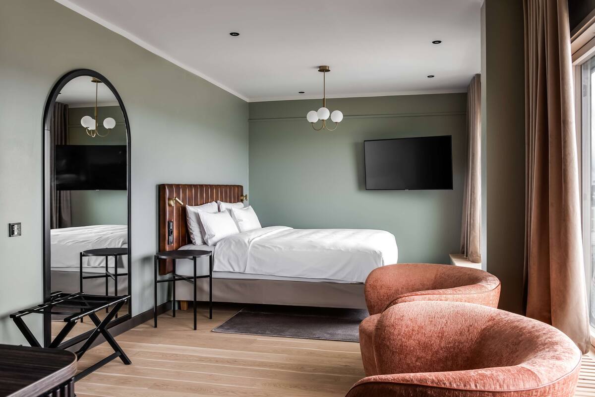 A modern hotel room with a bed, a desk, a chair, a TV, and a large mirror.