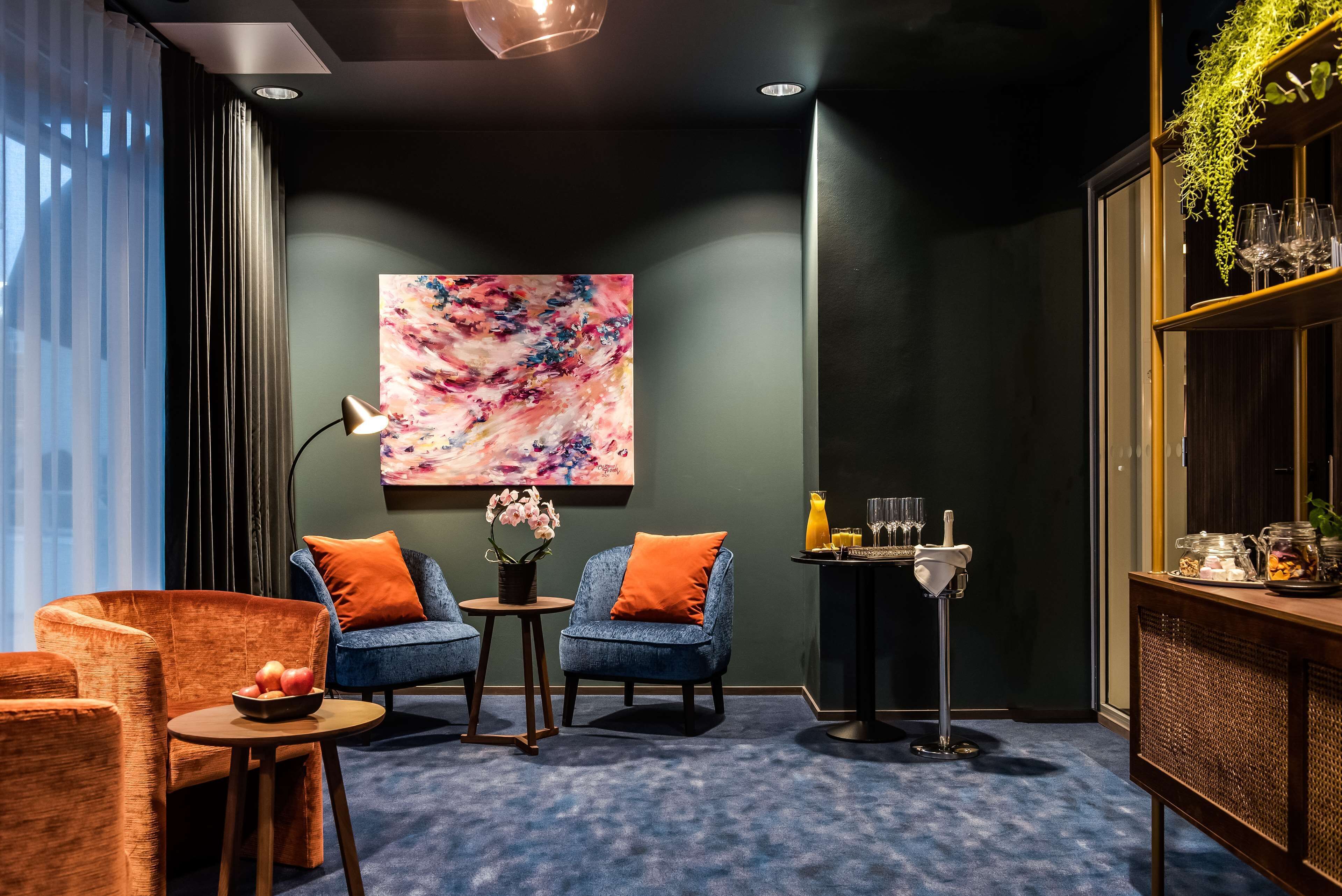 A modern lounge area with a blue carpet, two armchairs, a small table with a bowl of fruit, a floor lamp, and a wall-mounted painting.