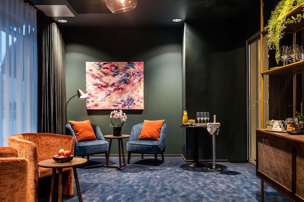 Un salon moderne avec un tapis bleu, deux fauteuils, une petite table sur laquelle se trouve un saladier de fruits, un lampadaire et un tableau accroché au mur.
