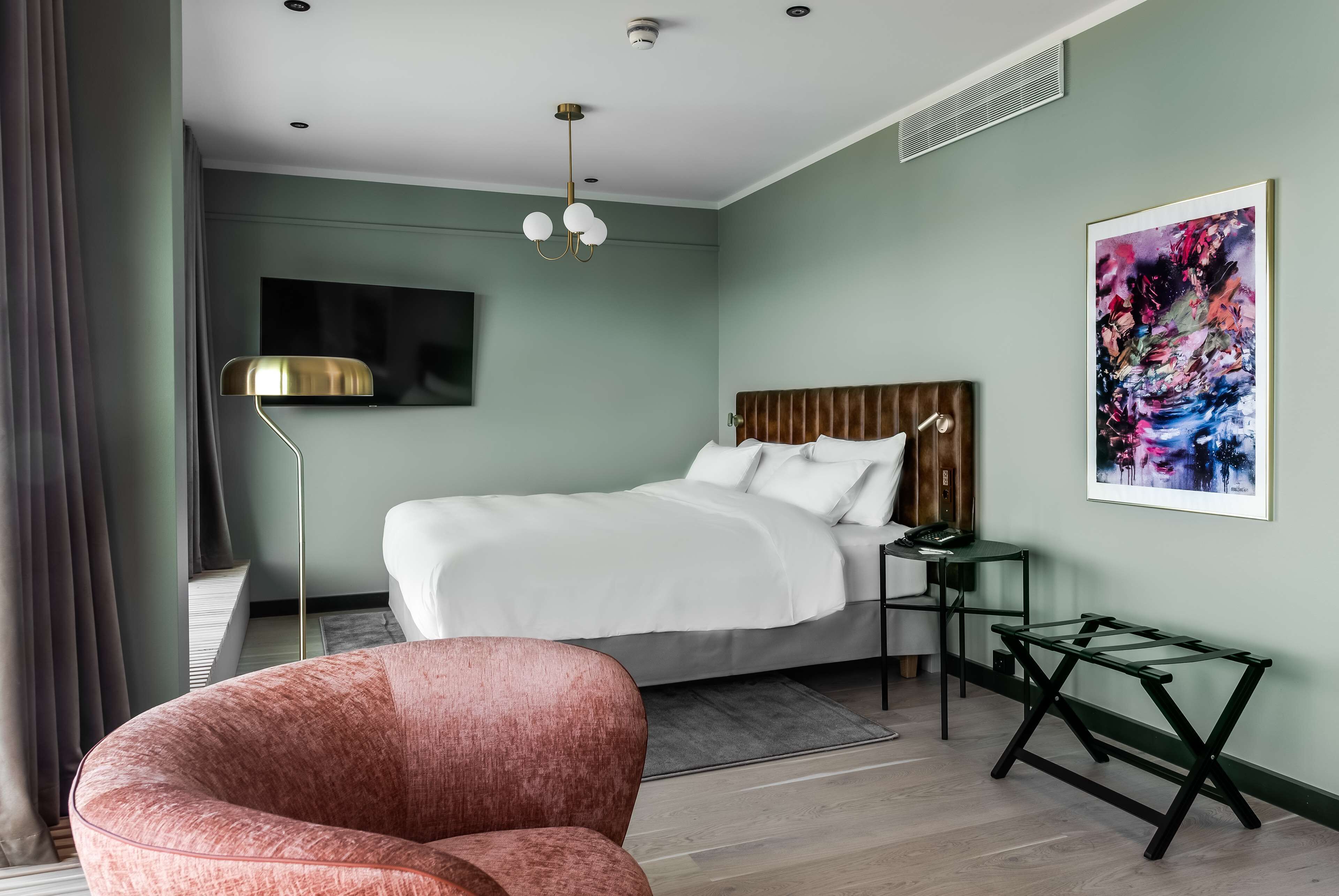 A modern hotel room with a large bed, a desk, a chair, a flat-screen TV, and a painting on the wall.