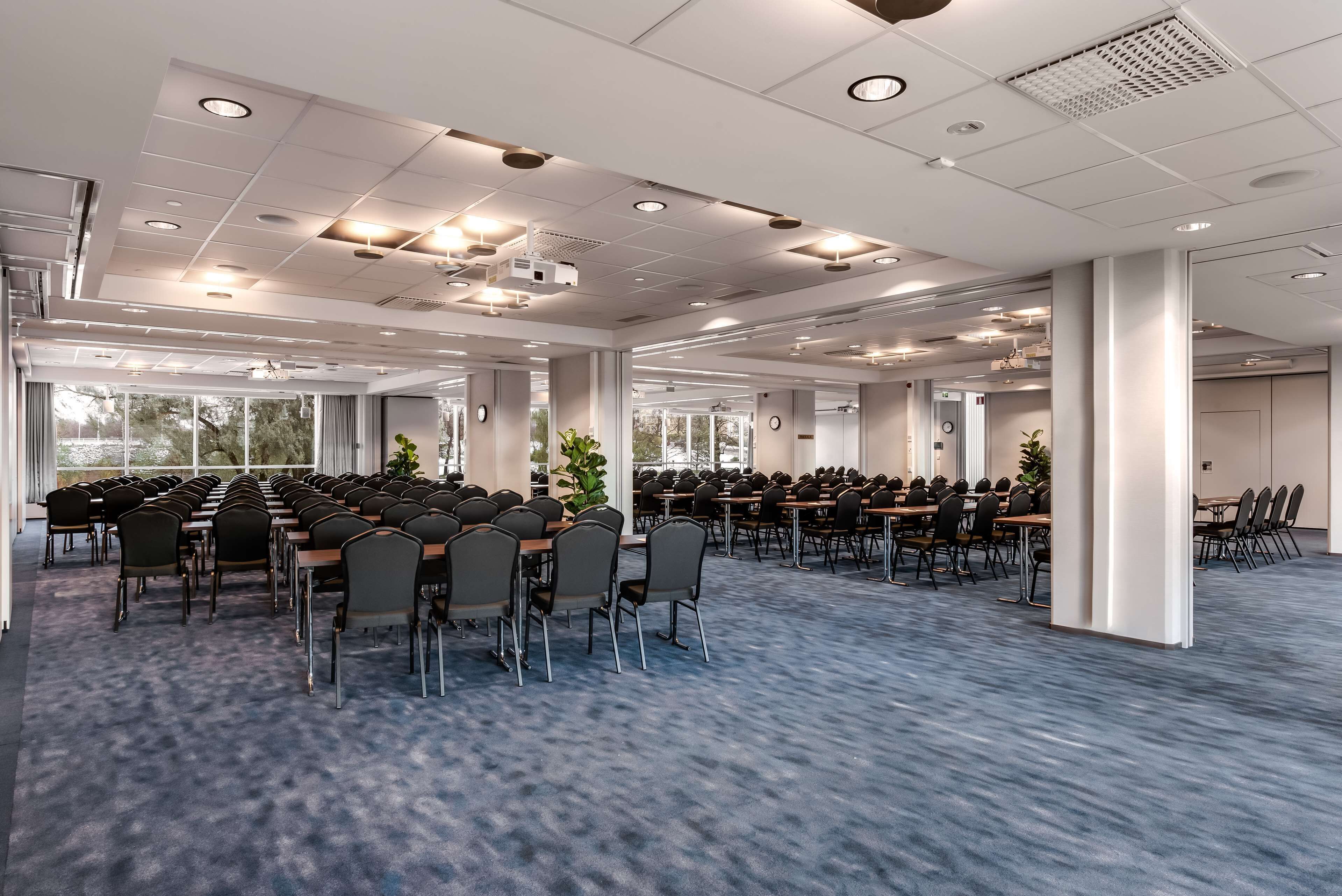 A large conference room with rows of tables and chairs, large windows, and a carpeted floor.