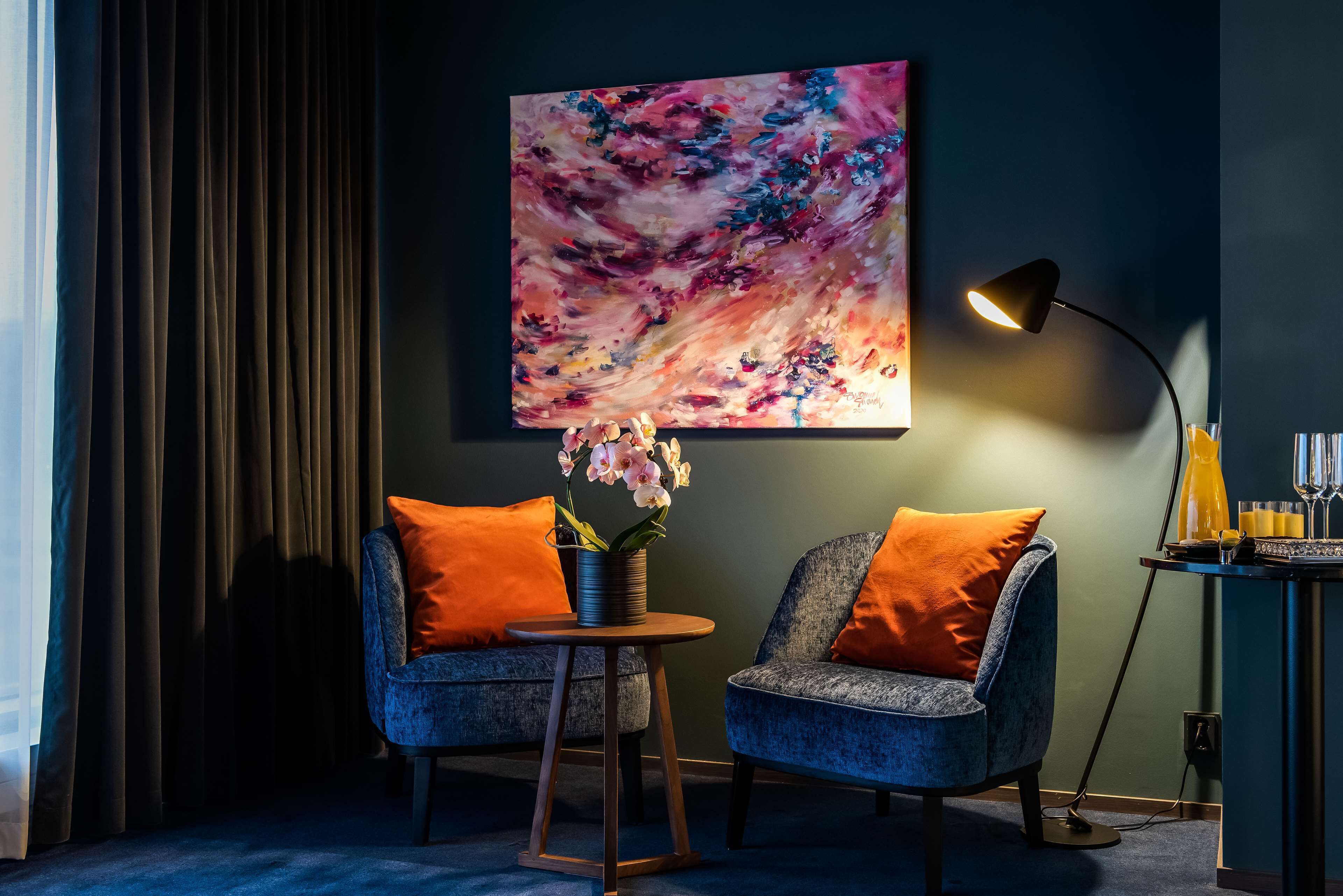 A room with two chairs, a table with a vase of flowers, a painting on the wall, and a lamp on a side table.
