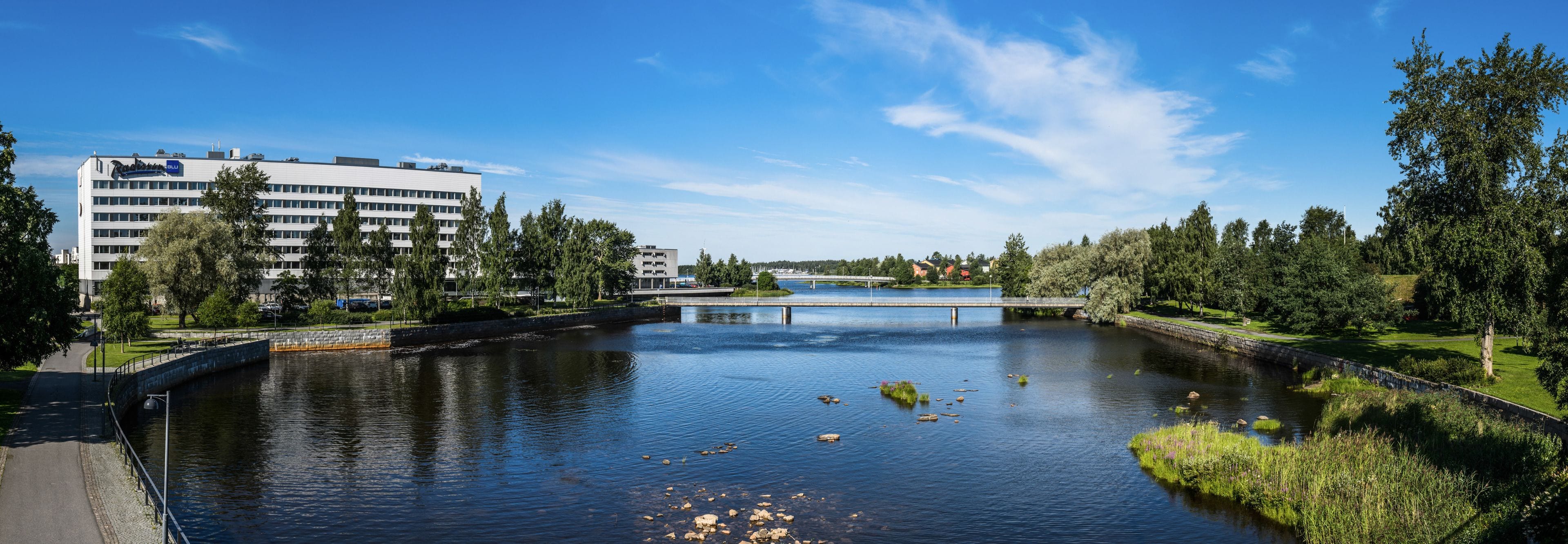 A river with a bridge, surrounded by greenery and a modern building with a sign.