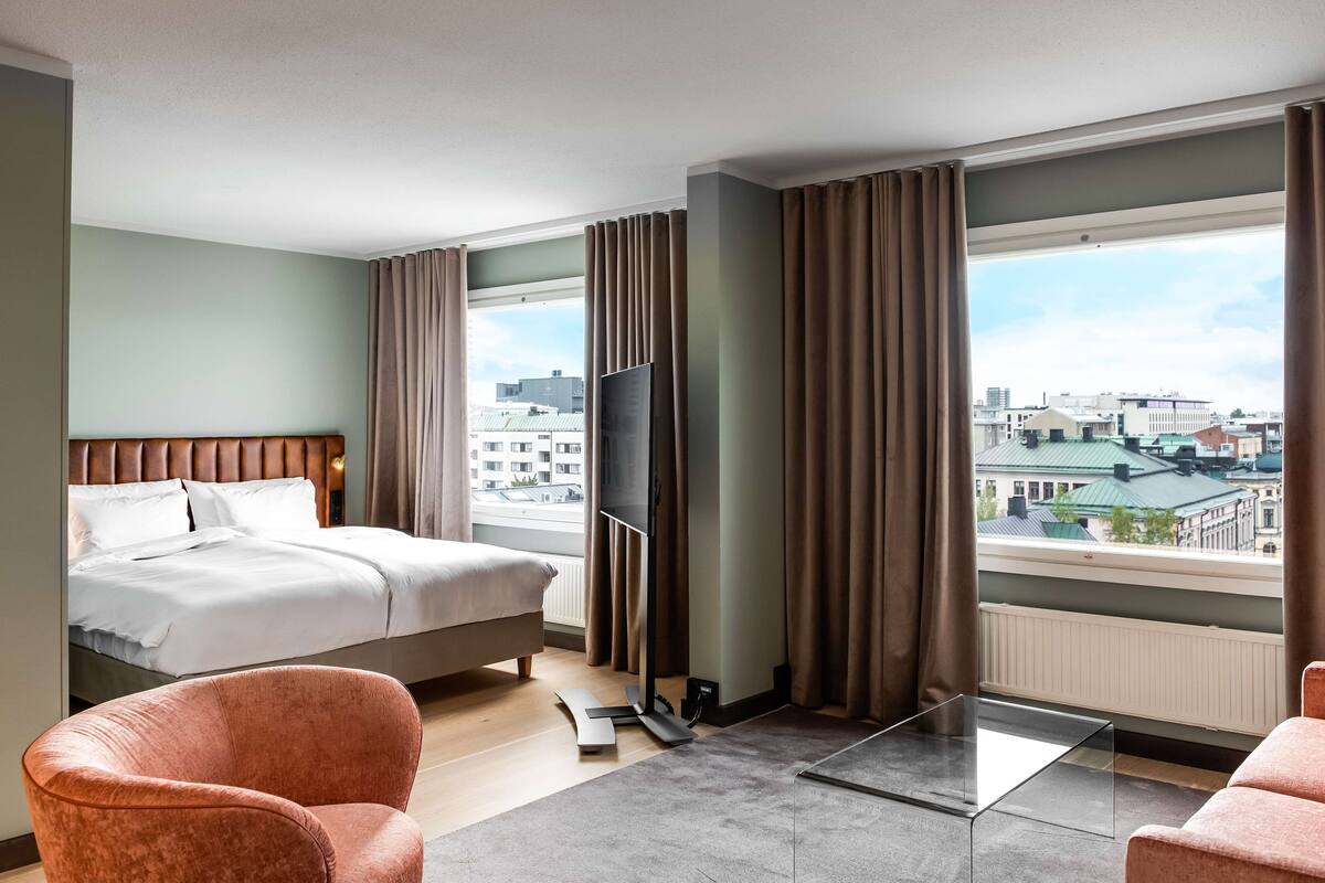 A hotel room with a bed, a TV, a glass table, and a view of the city.