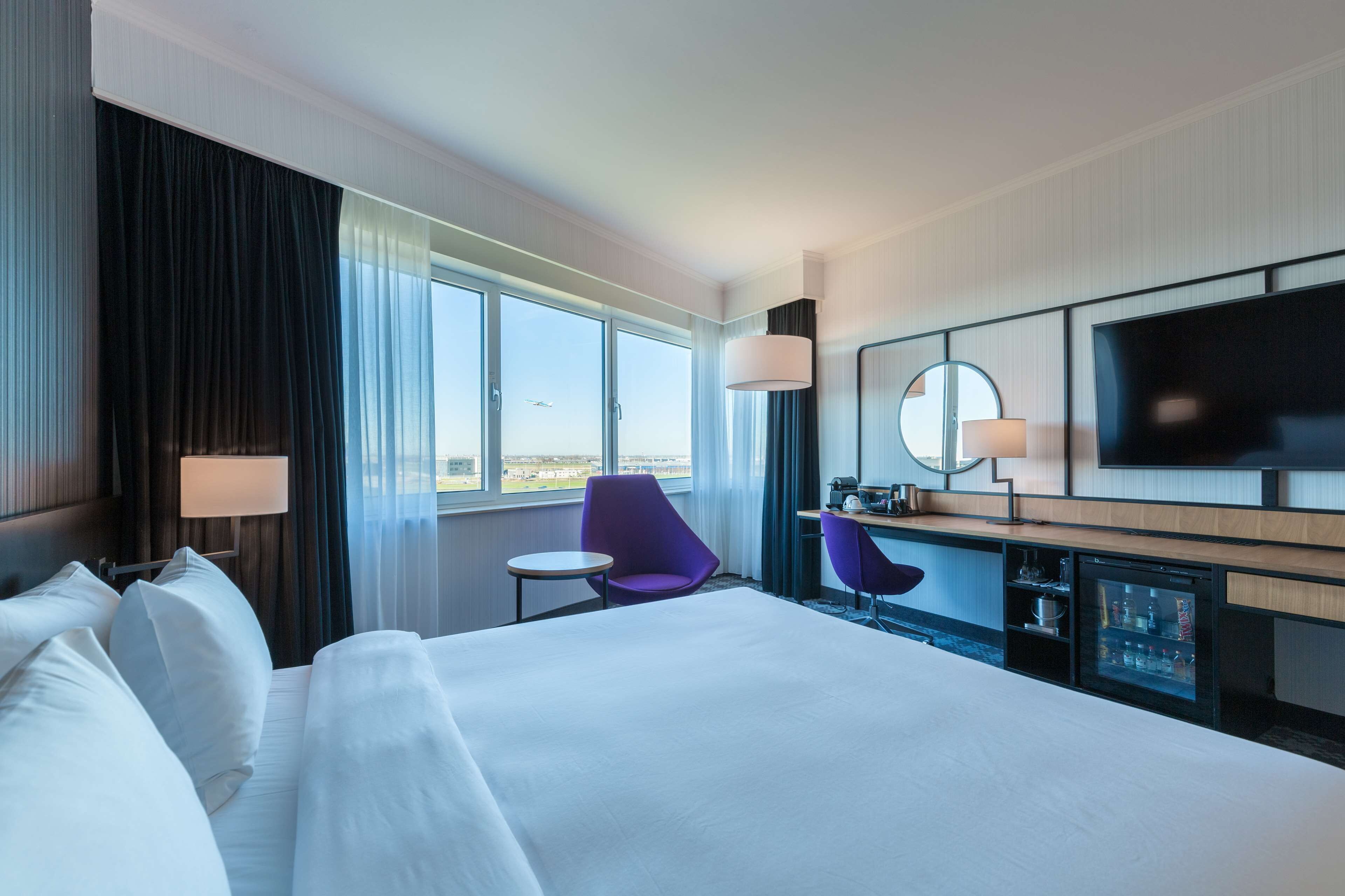 Ein Hotelzimmer mit einem großen Bett, einem Schreibtisch mit Computer, einem Fernseher und einem Fenster mit Blick auf den Himmel.