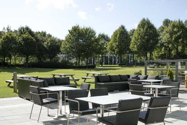 Ein Terrassenbereich im Freien mit mehreren Tischen und Stühlen, umgeben von Grünflächen und Bäumen.