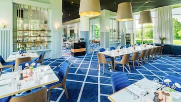 A restaurant with a blue carpet, tables set for dining, and large windows offering a view of the outdoors.