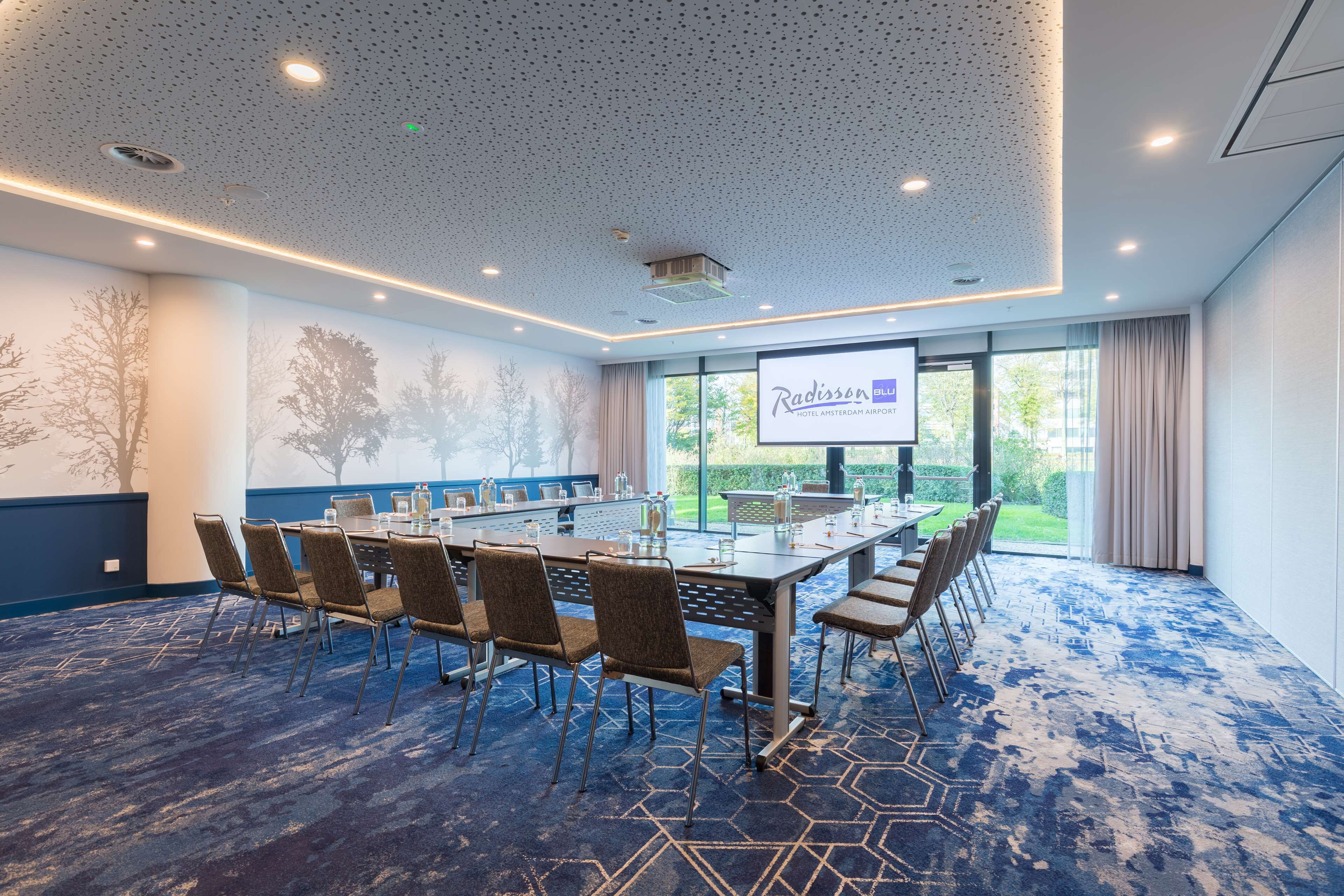 A conference room with a long table, chairs, and a projection screen displaying 'Radisson Blu Hotel Amsterdam Airport'.