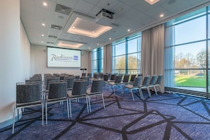 Une salle de conférence avec des rangées de chaises, un écran de projection affichant le logo de Radisson, et de grandes fenêtres donnant sur l’extérieur.