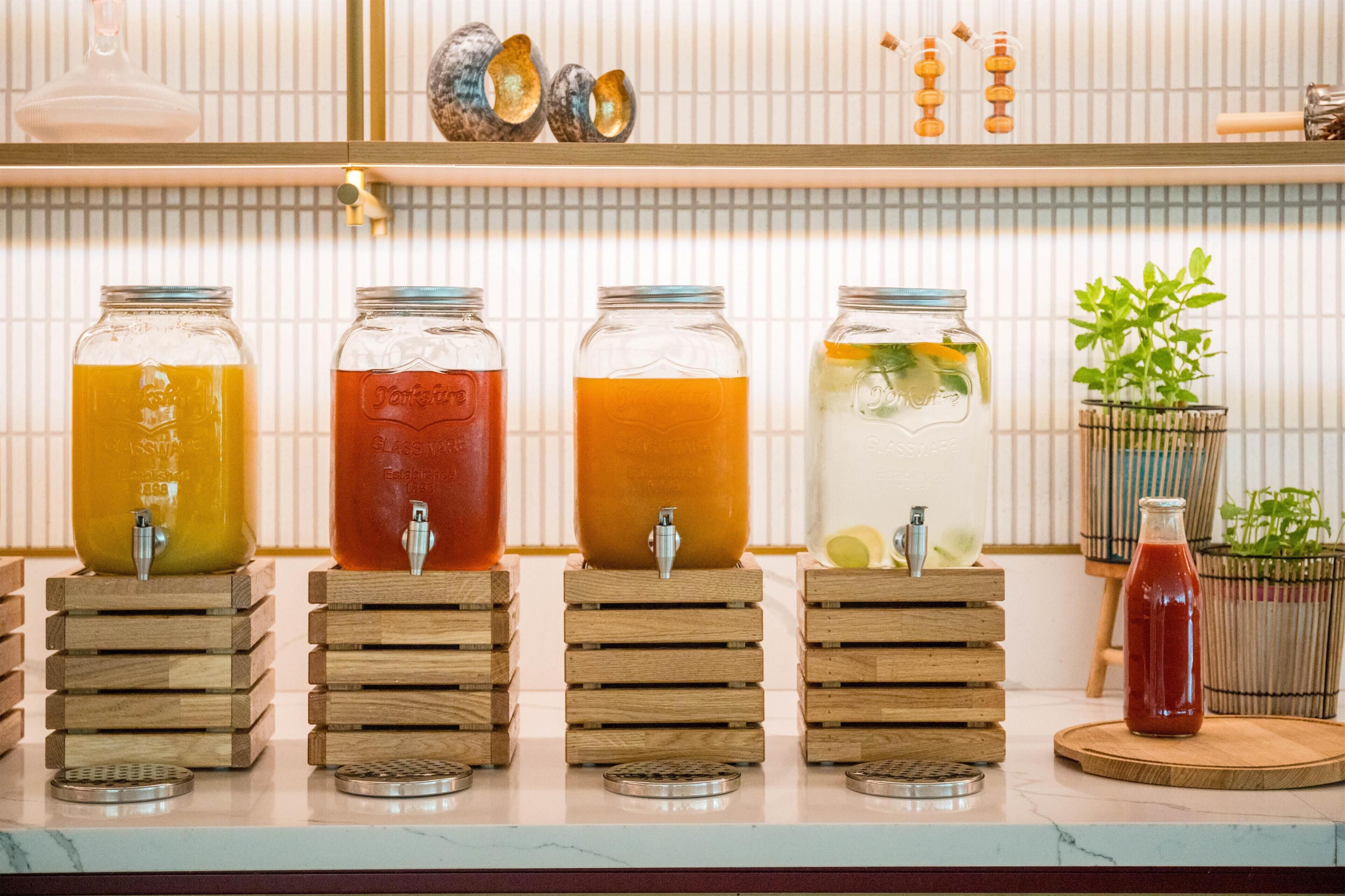 Cuatro soportes de madera con bebidas de diferentes colores, una pequeña planta en maceta y una botella de salsa roja sobre una encimera.