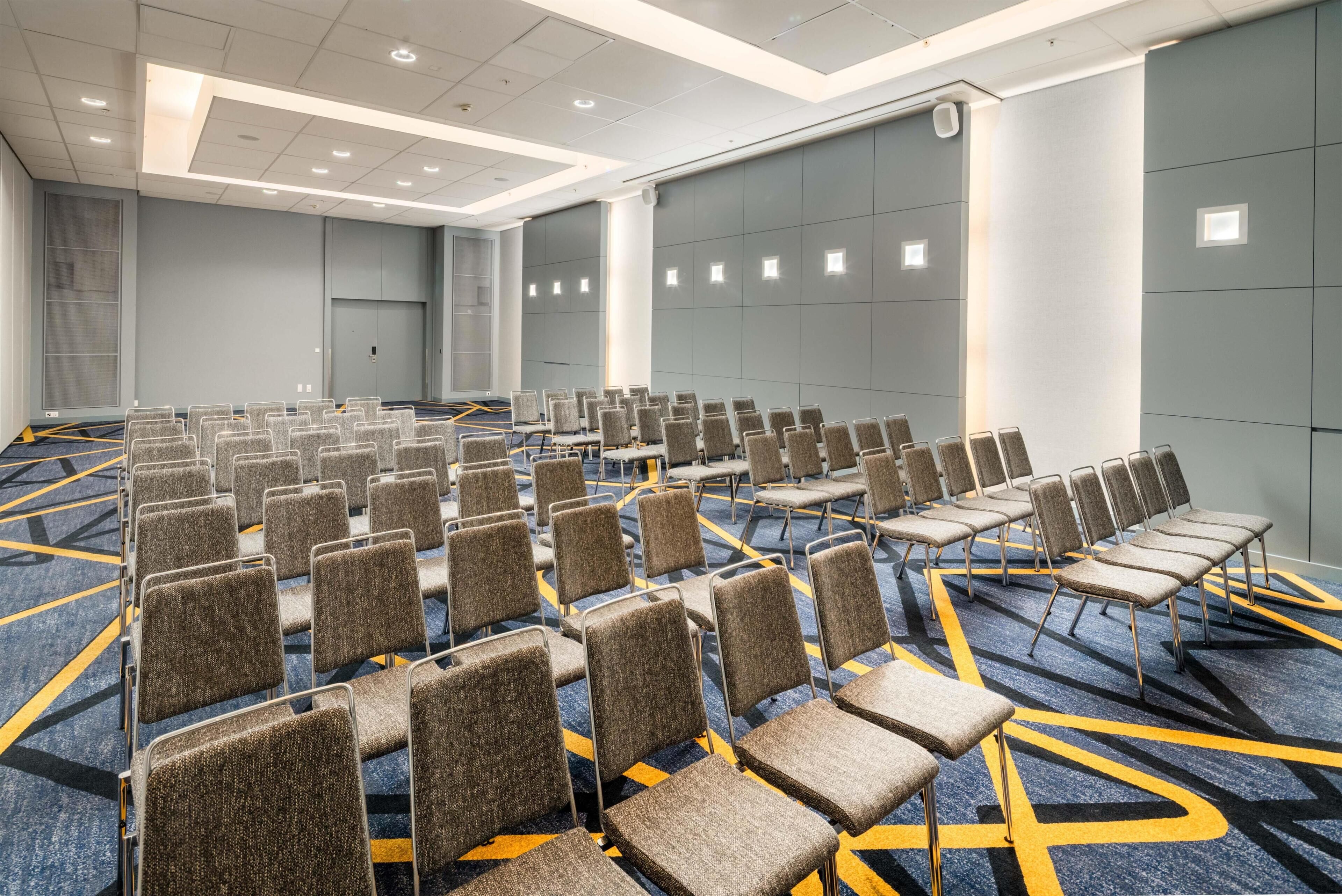 A conference room with rows of chairs, a carpeted floor, and a plain wall with a door.