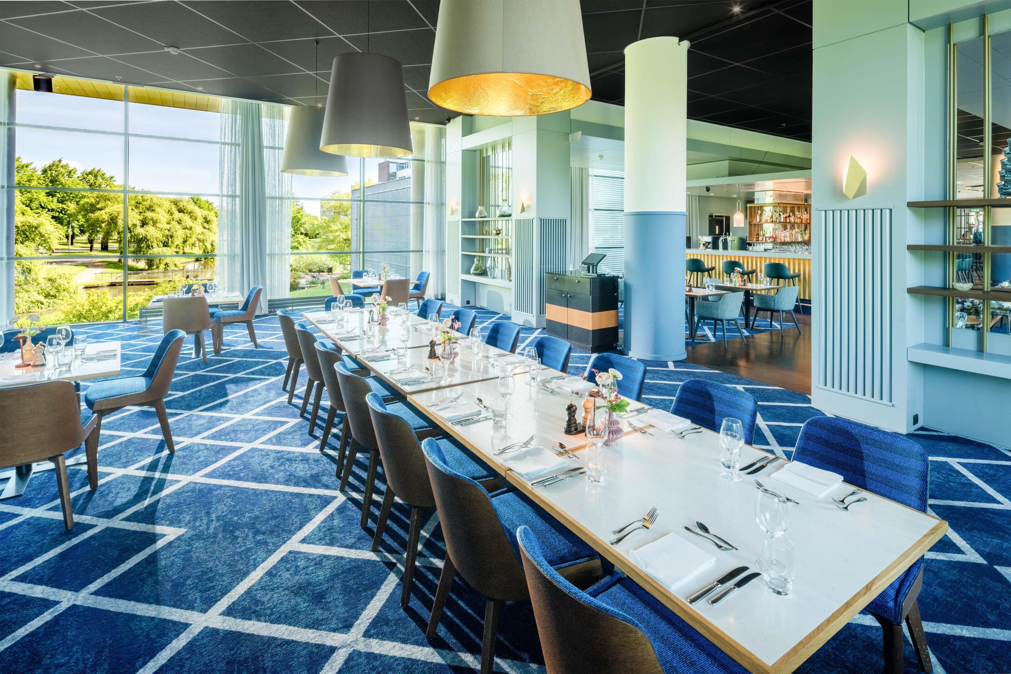 Um restaurante com uma mesa comprida posta para refeições, grandes janelas e vista para o exterior.
