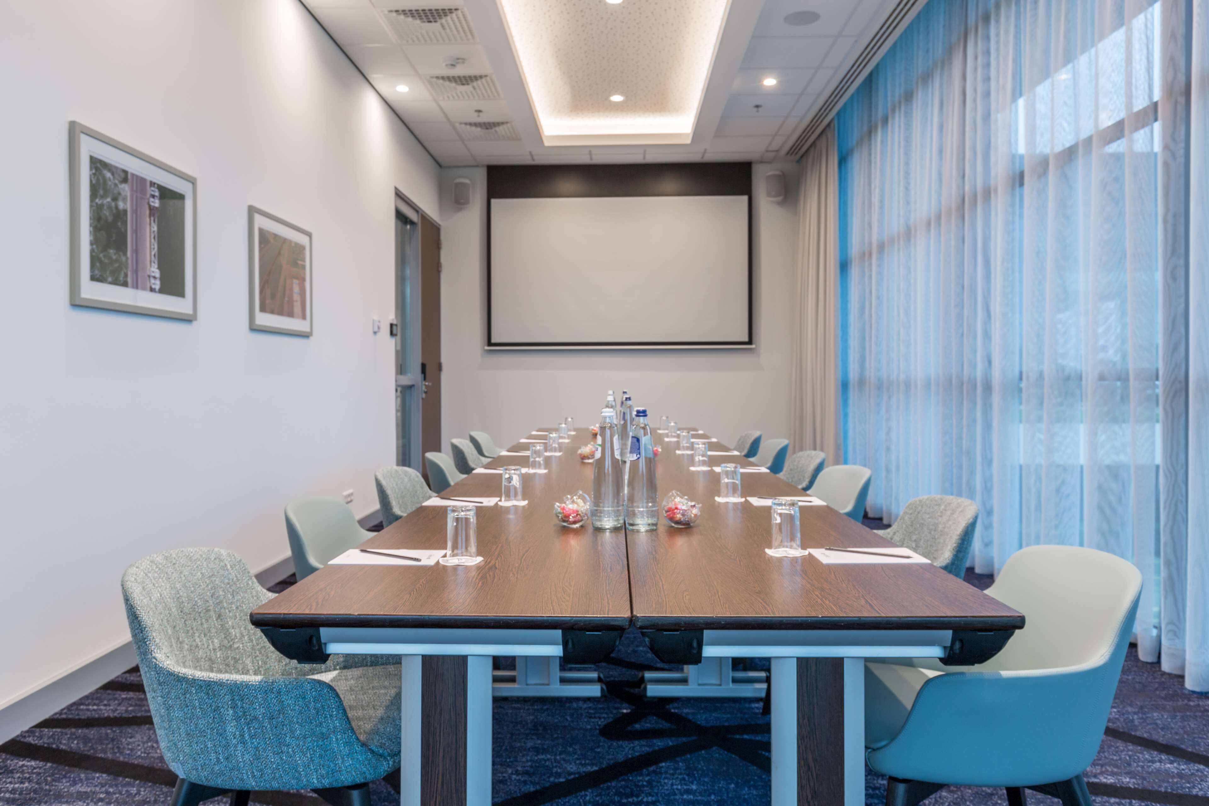 Une salle de conférence équipée d’une longue table, de chaises, d’un écran de projection et d’une grande fenêtre avec des rideaux.