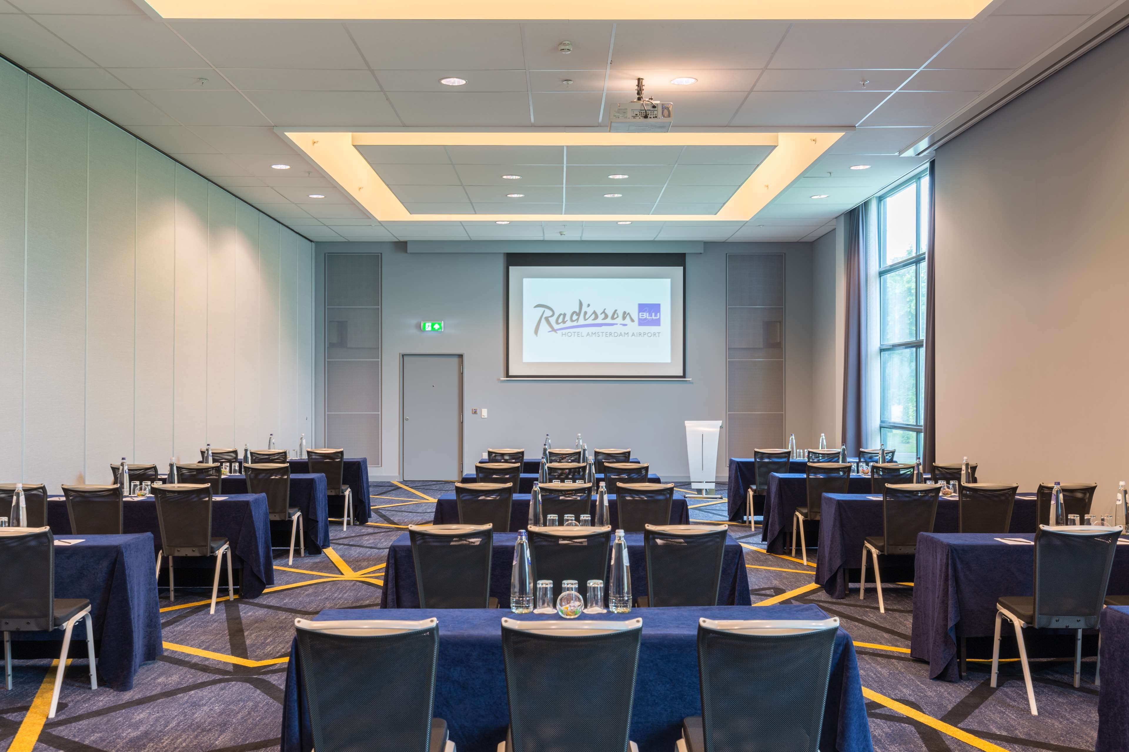 A conference room with multiple tables and chairs, a projector screen displaying a presentation, and a large window providing natural light.