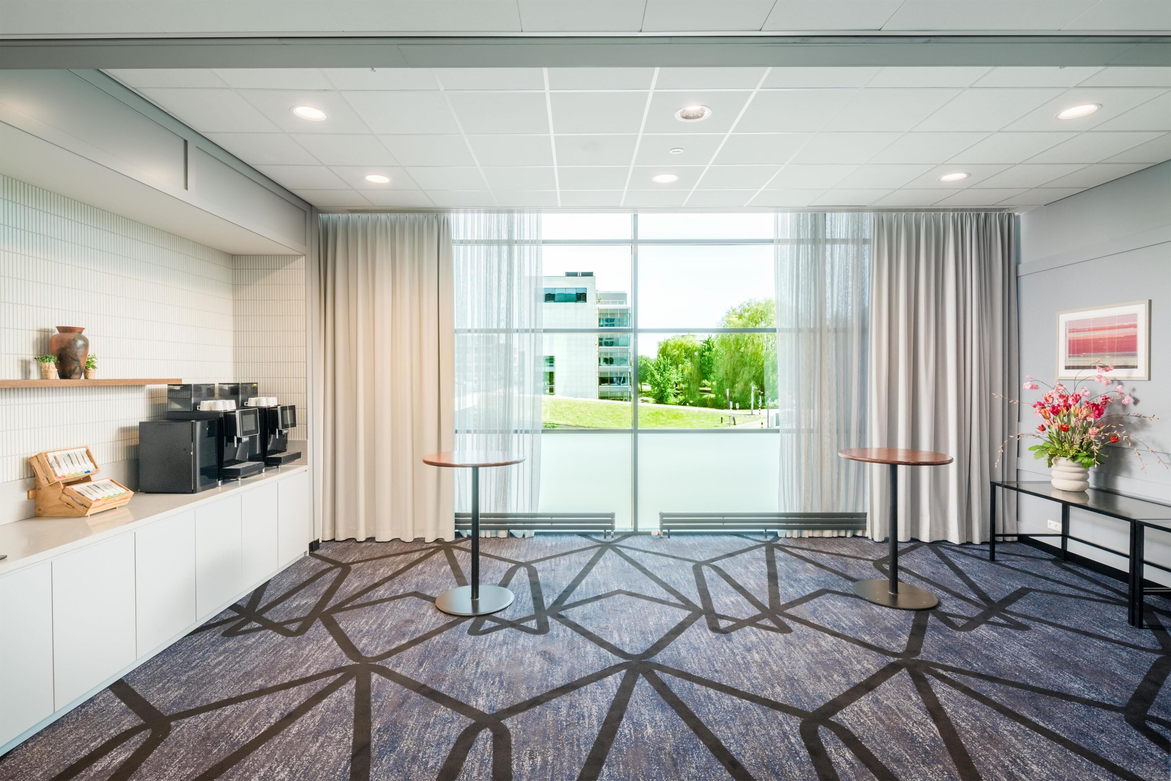 Un vestíbulo de hotel con una recepción, máquinas de café, una zona de estar con mesas redondas y ventanas amplias que ofrecen vistas al exterior.