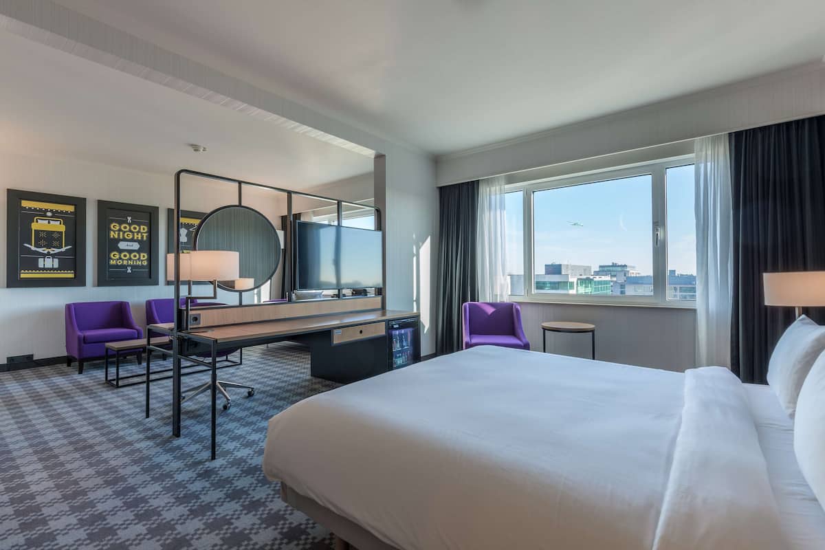Ein Hotelzimmer mit einem großen Bett, einem Schreibtisch mit Stuhl, einem kleinen Tisch und einem großen Fenster mit Blick auf die Stadt.