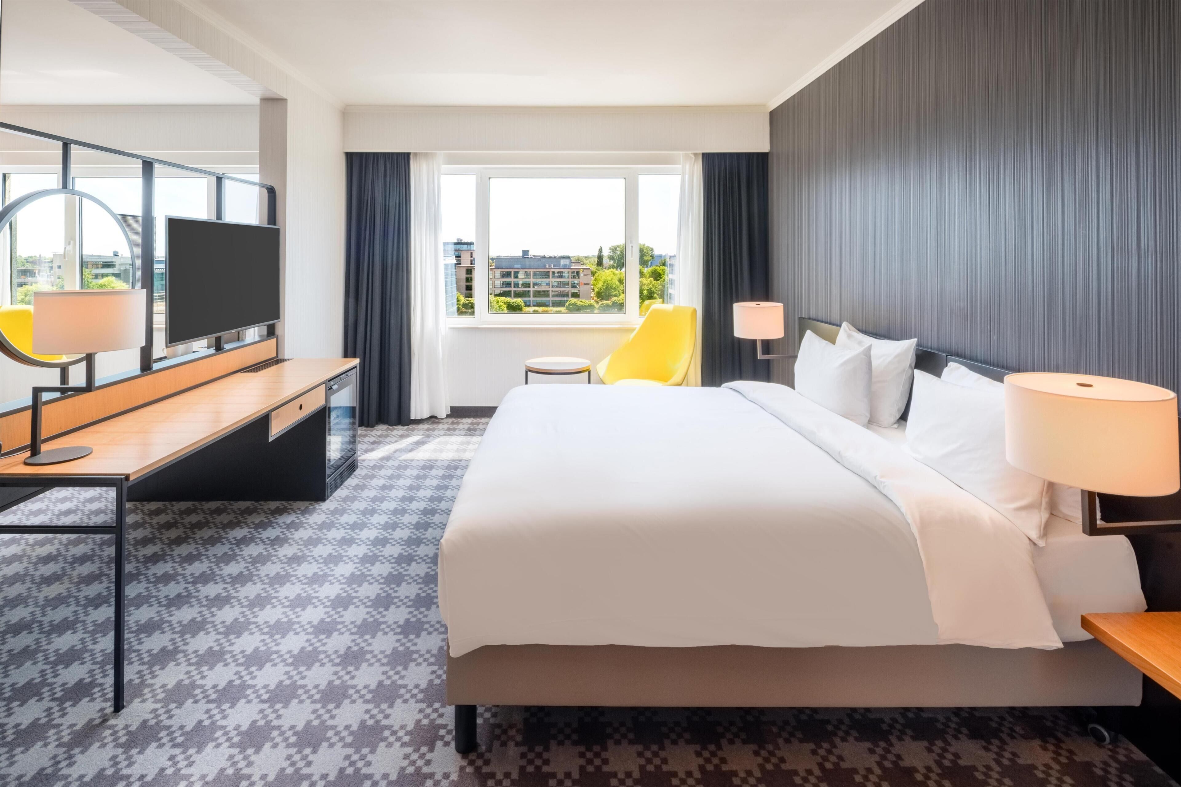 A hotel room with a bed, a desk with a TV, a chair, a window with a view, and a lamp.