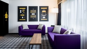 Ein modernes Hotelzimmer mit einem violetten Sofagarnitur, einem Holztisch und drei gerahmten Kunstwerken an der Wand.