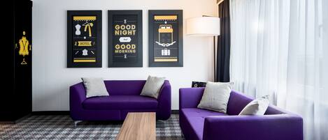 A modern hotel room with a purple sofa set, a wooden coffee table, and three framed wall art pieces.