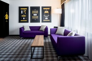 Ein modernes Hotelzimmer mit einem violetten Sofagarnitur, einem Holztisch und drei gerahmten Kunstwerken an der Wand.