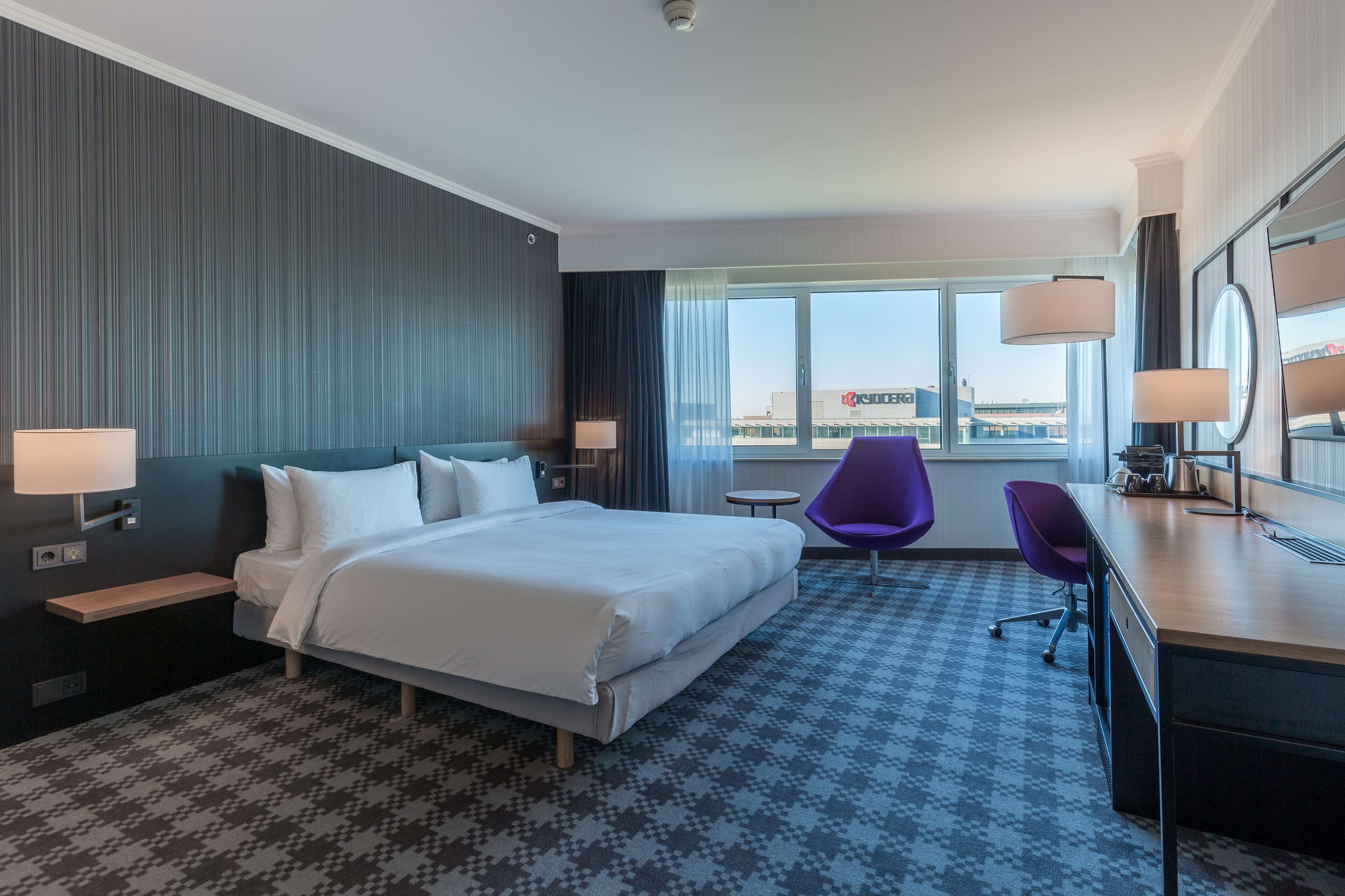 A hotel room with a large bed, a desk with a chair, a small round table, and a view of the city through the window.