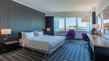 A hotel room with a large bed, a desk with a chair, a small round table, and a view of the city through the window.