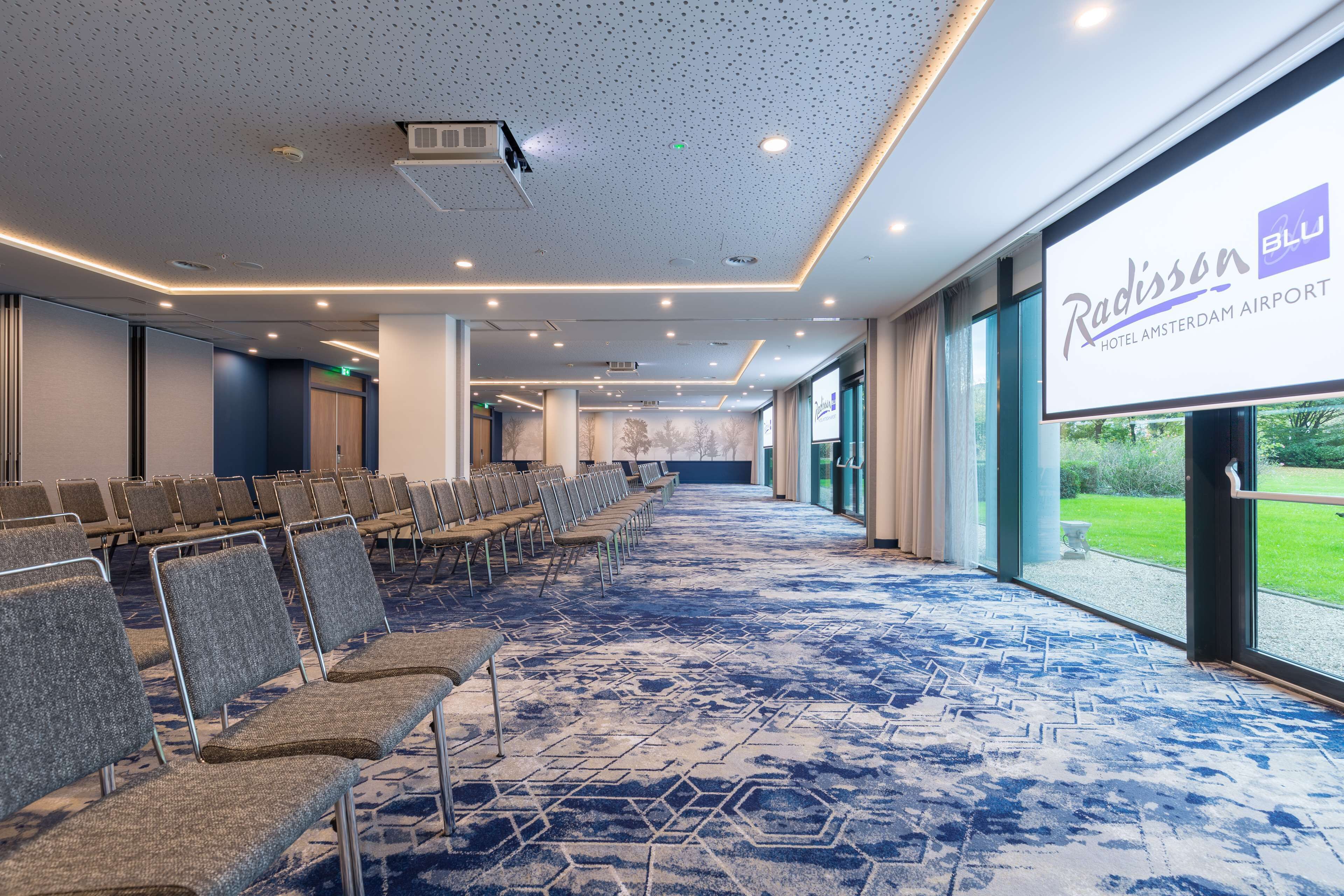 Un salón de conferencias con filas de sillas, una pantalla de proyector y vistas al aeropuerto.