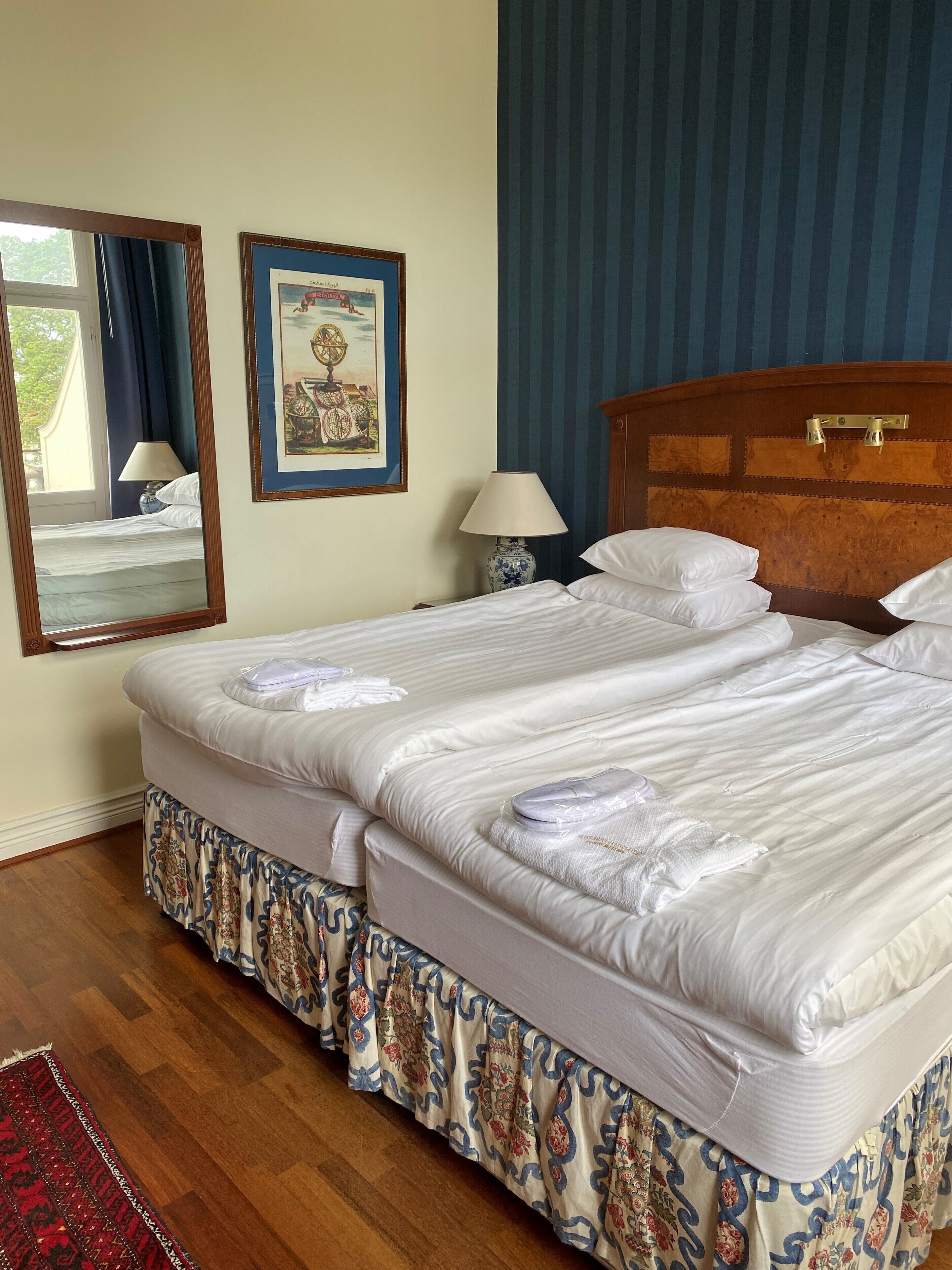 A hotel room with two beds, a wooden headboard, a framed picture on the wall, a bedside lamp, and a red patterned rug.