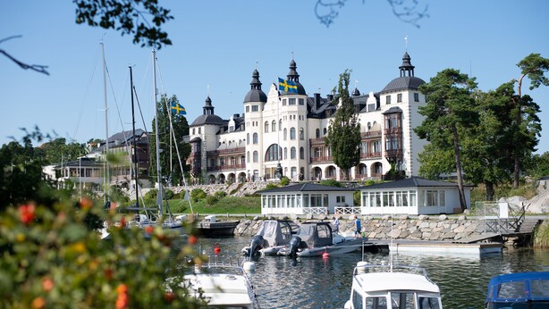 Ein großes, historisches Gebäude mit mehreren Kuppeln und Flaggen, gelegen an einem Gewässer mit Booten im Hafen.