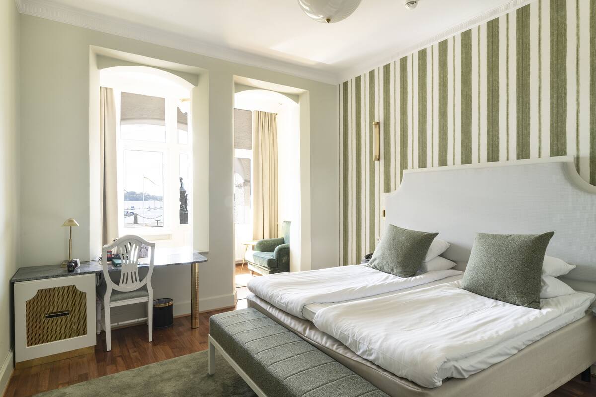 Ein Schlafzimmer mit einem großen Bett, einem Schreibtisch und einem Stuhl. Zwei Fenster mit Vorhängen und ein Blick ins Freie.