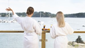 Zwei Personen in weißen Roben blicken auf einen Yachthafen mit Booten und einer felsigen Küstenlinie.