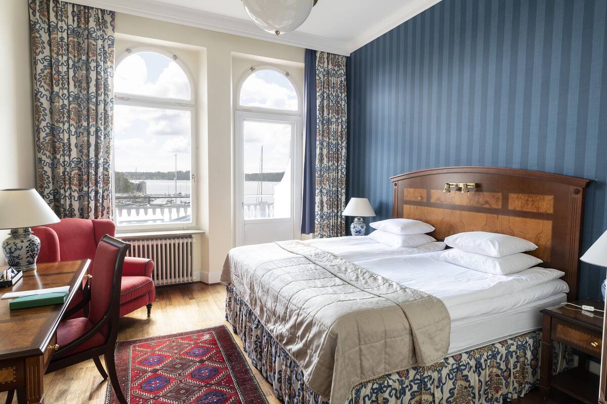 Ein Hotelzimmer mit einem großen Bett, zwei Vorhängen, einem roten Sessel, einem Holztisch mit einer Lampe und Blick auf das Wasser.