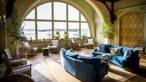 Ein geräumiges Wohnzimmer mit einem großen Bogenfenster mit Blick auf ein Gewässer, möbliert mit blauen Sofas, Korbstühlen und Topfpflanzen.