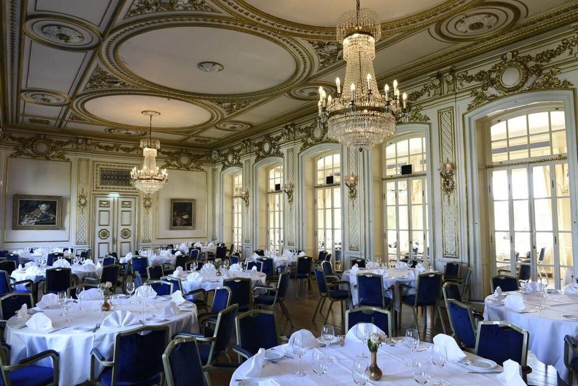 Un salón comedor majestuoso con candelabros elaborados, amplias ventanas y mesas elegantemente dispuestas.