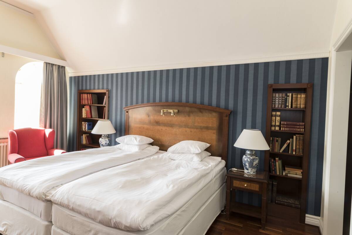 Ein Hotelzimmer mit einem großen Bett, zwei Nachttischen mit Lampen, einem roten Sessel und zwei Bücherregalen voller Bücher.