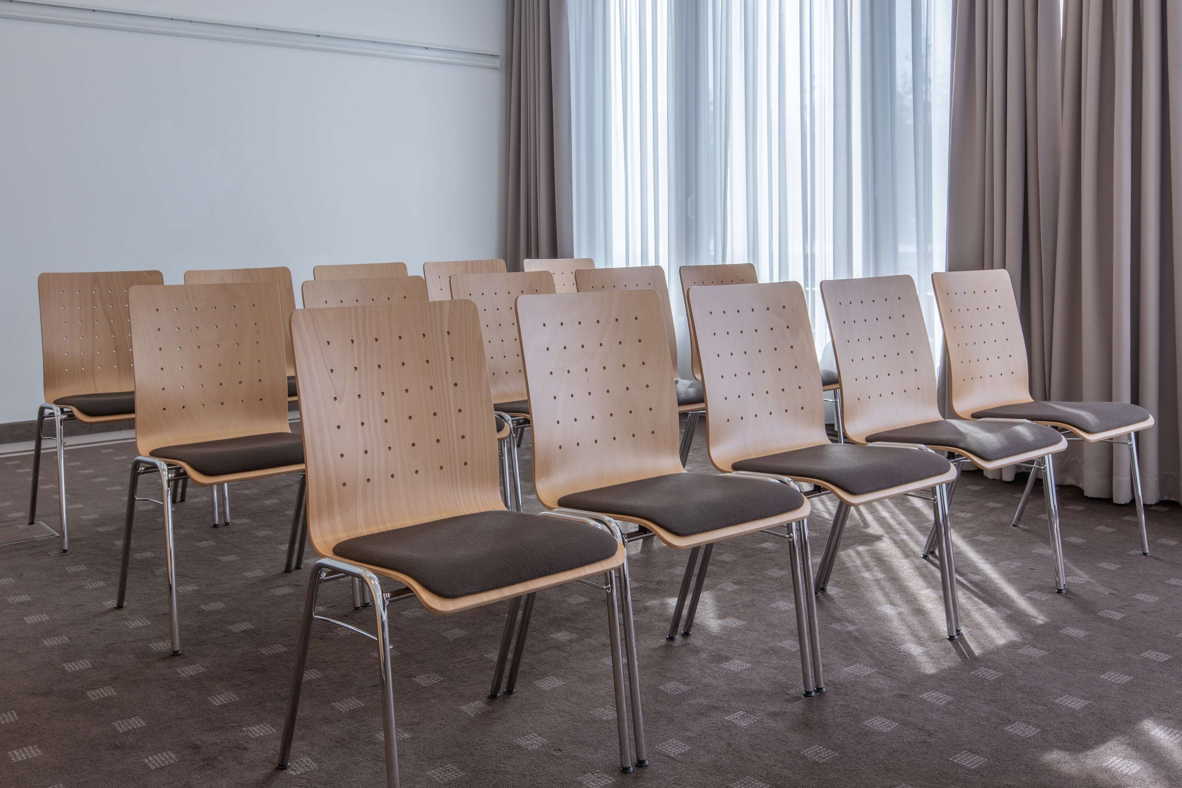 Eine Reihe Holzstühle mit Metallbeinen und gepolsterten Sitzflächen, angeordnet in einem Konferenzraum mit Teppichboden und einem fenster mit Vorhängen.