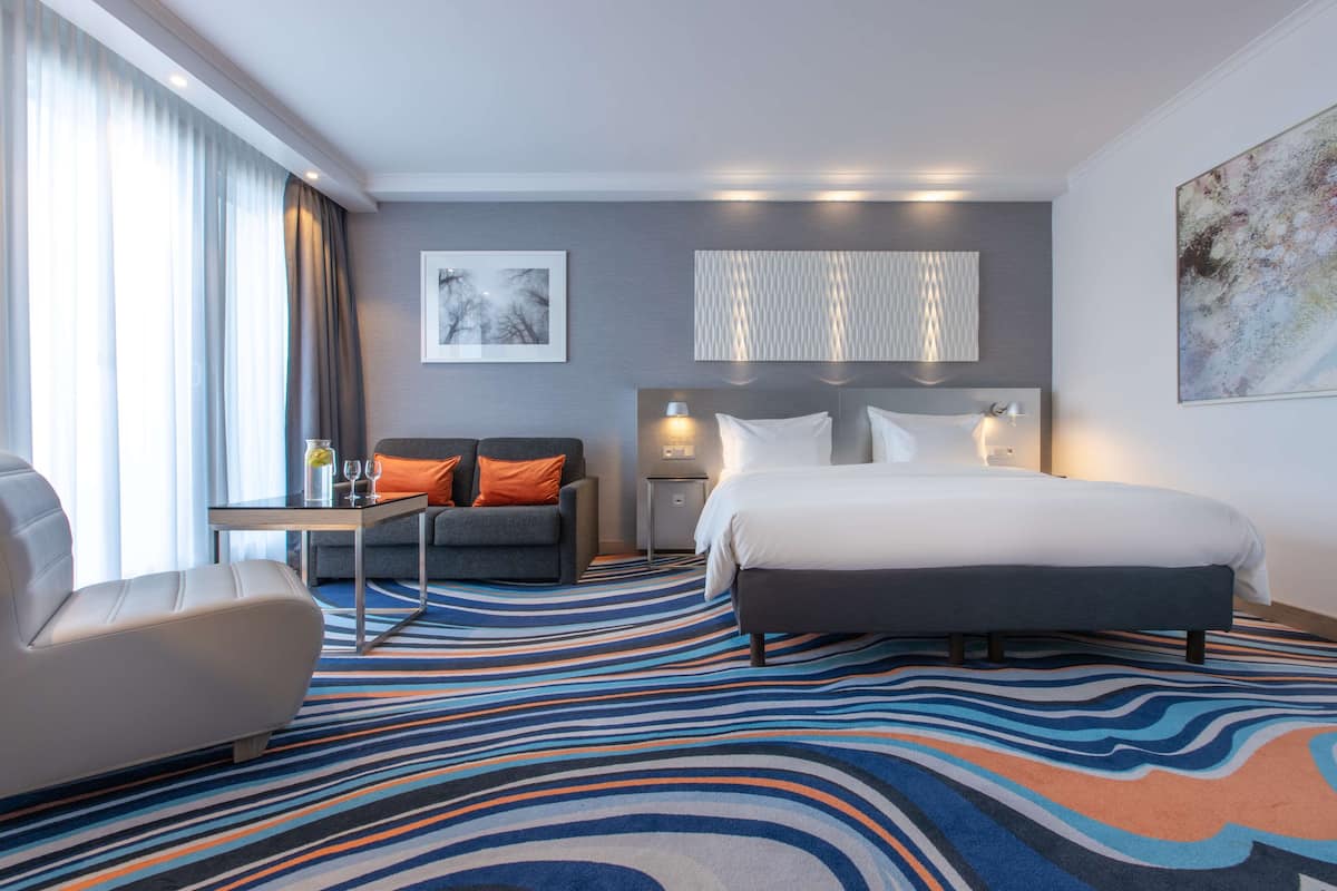 A hotel room with a large bed, a sofa, a small table with glasses, and a view of the outdoors through large windows.