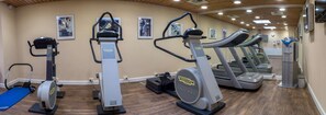 Un gimnasio con diversas máquinas de cardio, incluyendo elípticas y cintas de correr, dispuesto en una sala amplia con piso de madera y paredes color beige.