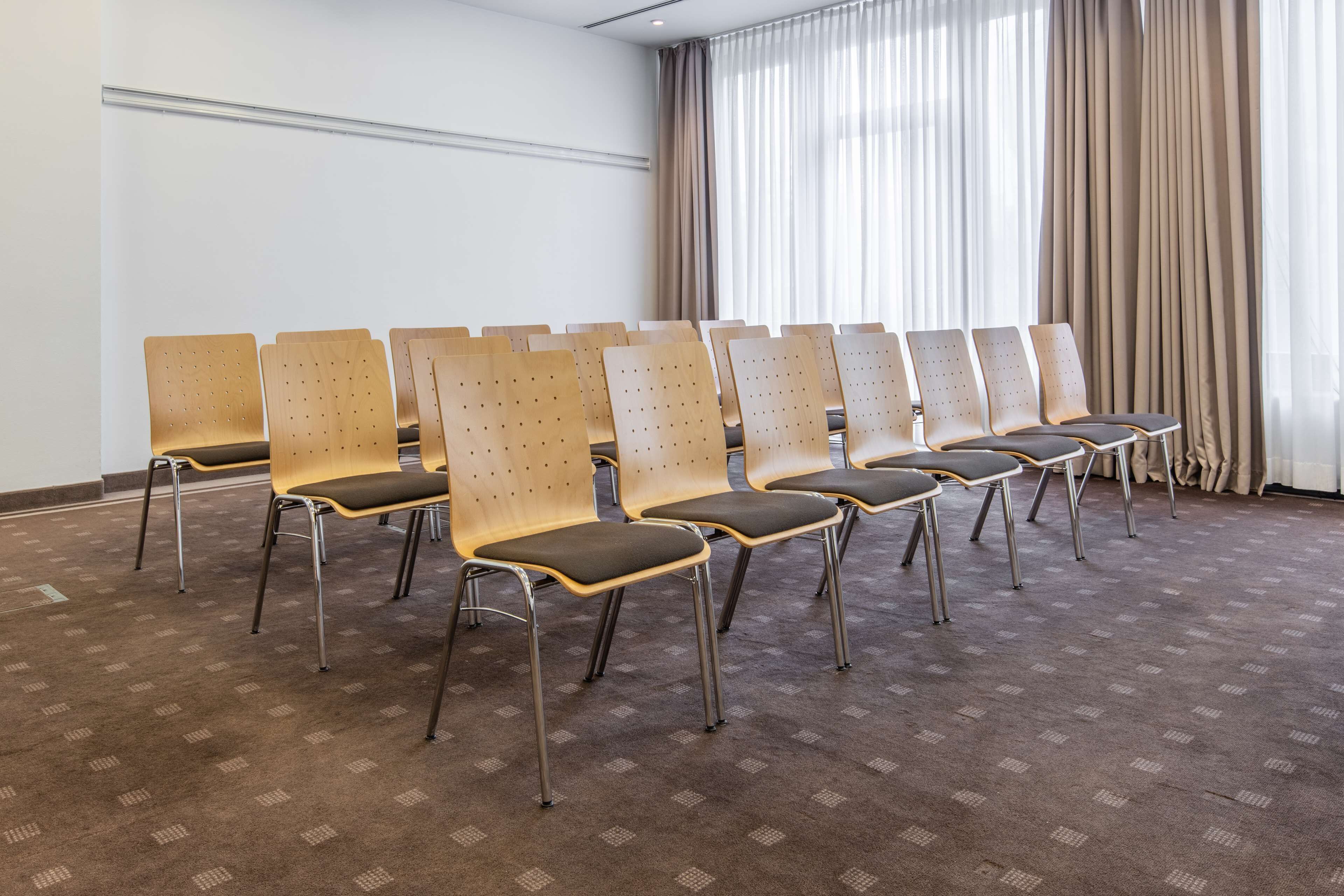 A conference room with rows of wooden chairs with perforated backs and cushioned seats, arranged on a patterned carpet.