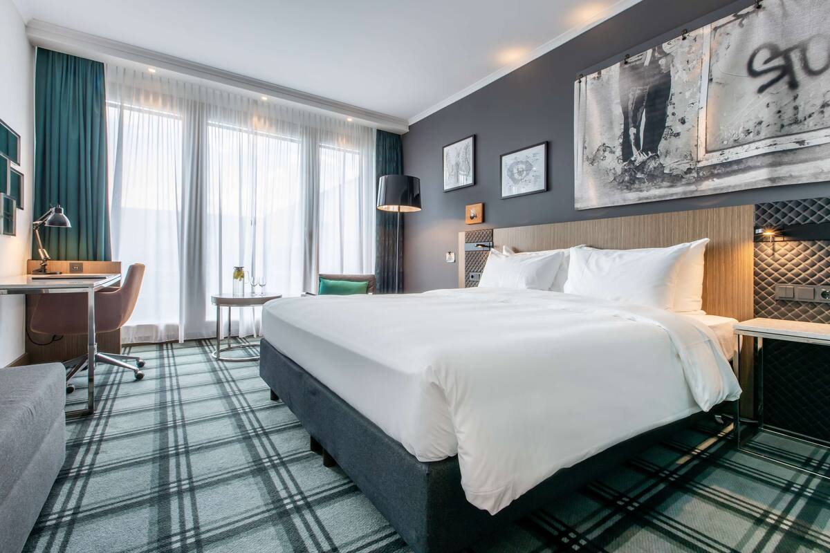 A hotel room with a large bed, a desk, a chair, and a small table. There are curtains on the windows, and artwork on the walls.