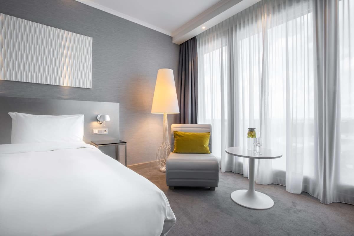 A modern hotel room with a large bed, a seating area with a lamp, and a view through sheer curtains.
