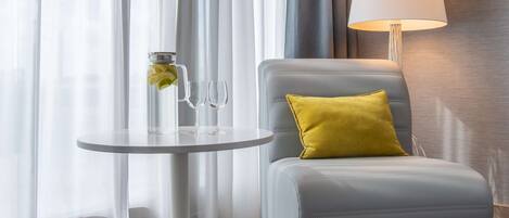 Ein modernes Zimmer mit einem grauen Sessel, einem kleinen runden Tisch mit einem Glasbehälter und zwei Gläsern, einem gelben Kissen, einer weißen Lampe und leichten Vorhängen.